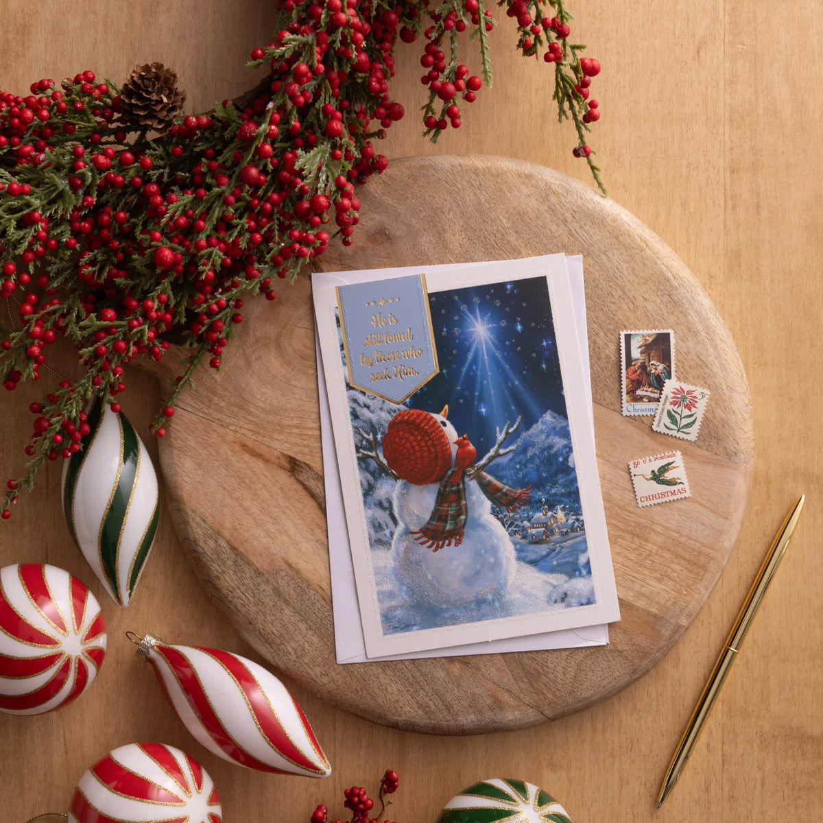 Snowman and star Christmas card on a wooden board with berry garland, ornaments and stamps