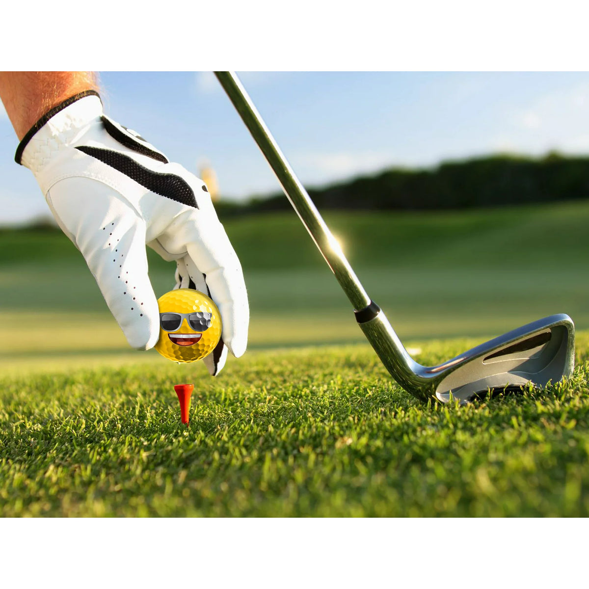 Gloved hand placing a sunglasses emoji golf ball on a tee beside an iron