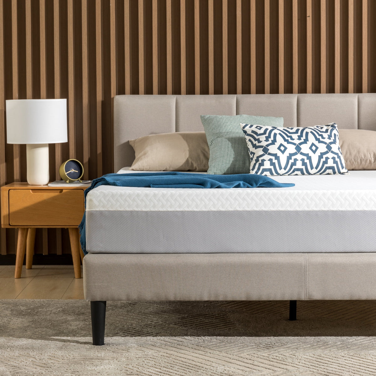 Mattress on an upholstered bed with a teal throw, pillows and a wooden nightstand lamp