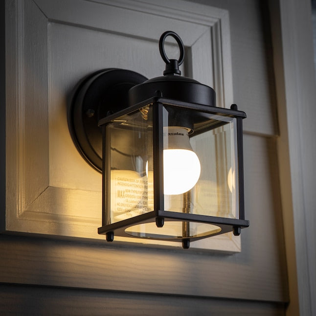 Lit black lantern wall light glowing on a grey-sided house at dusk