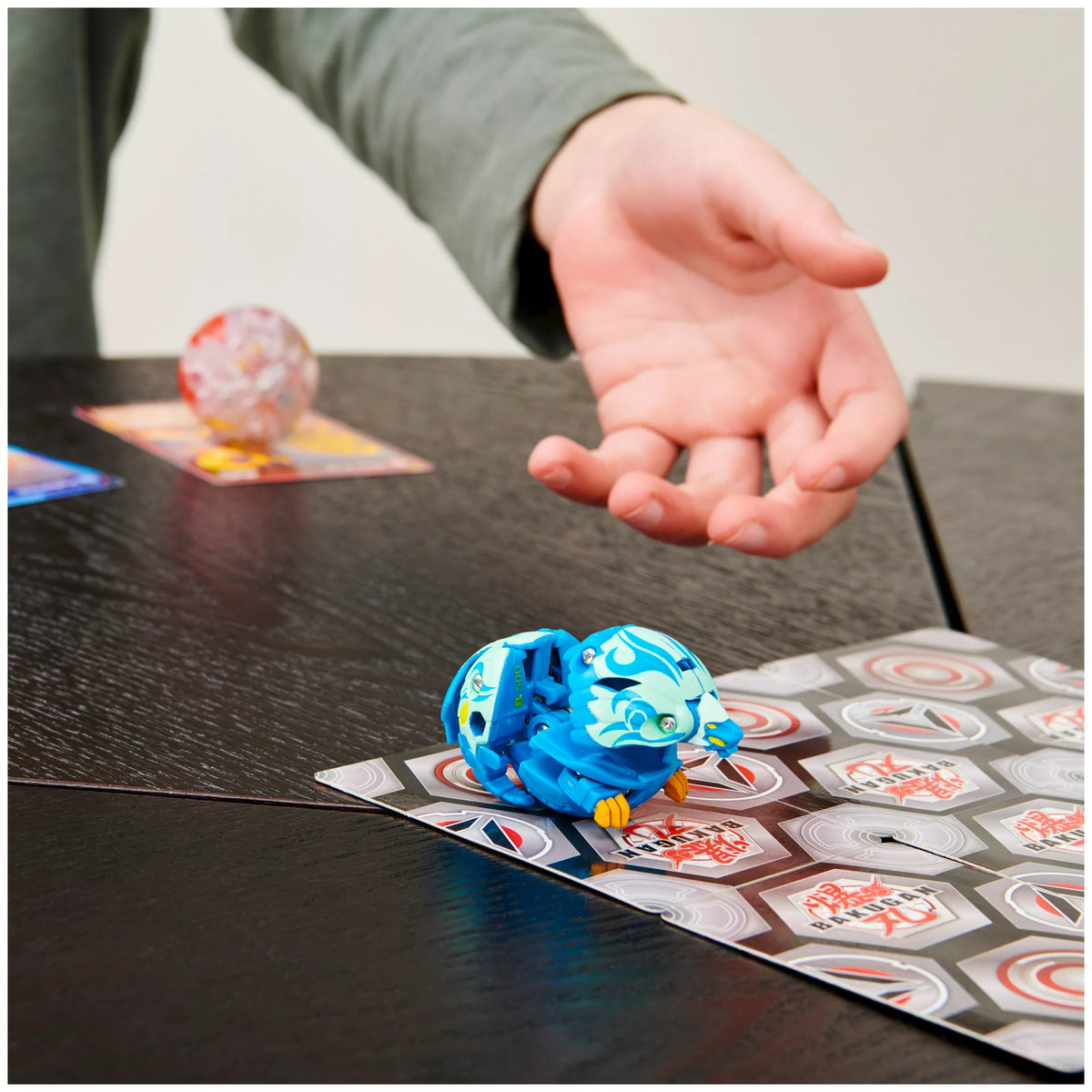 Hand rolling a blue Bakugan that pops open onto a hex-pattern game mat