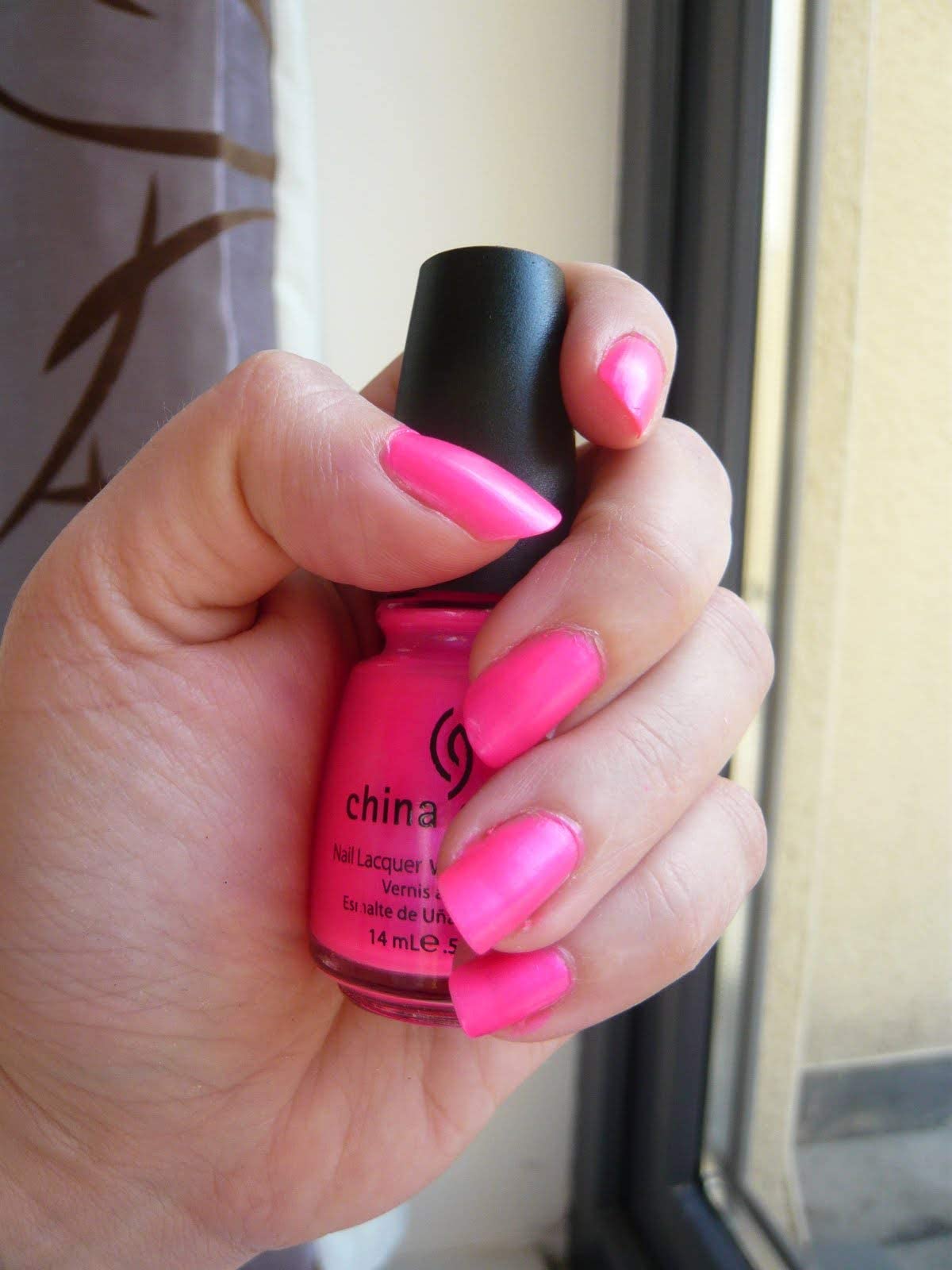 Hand with long neon pink nails holding the polish bottle by a window