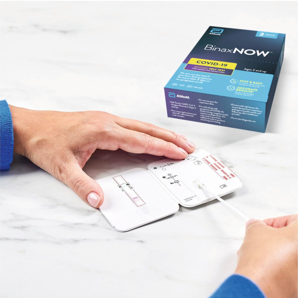 Hands inserting a nasal swab into an open BinaxNOW test card on marble, box behind