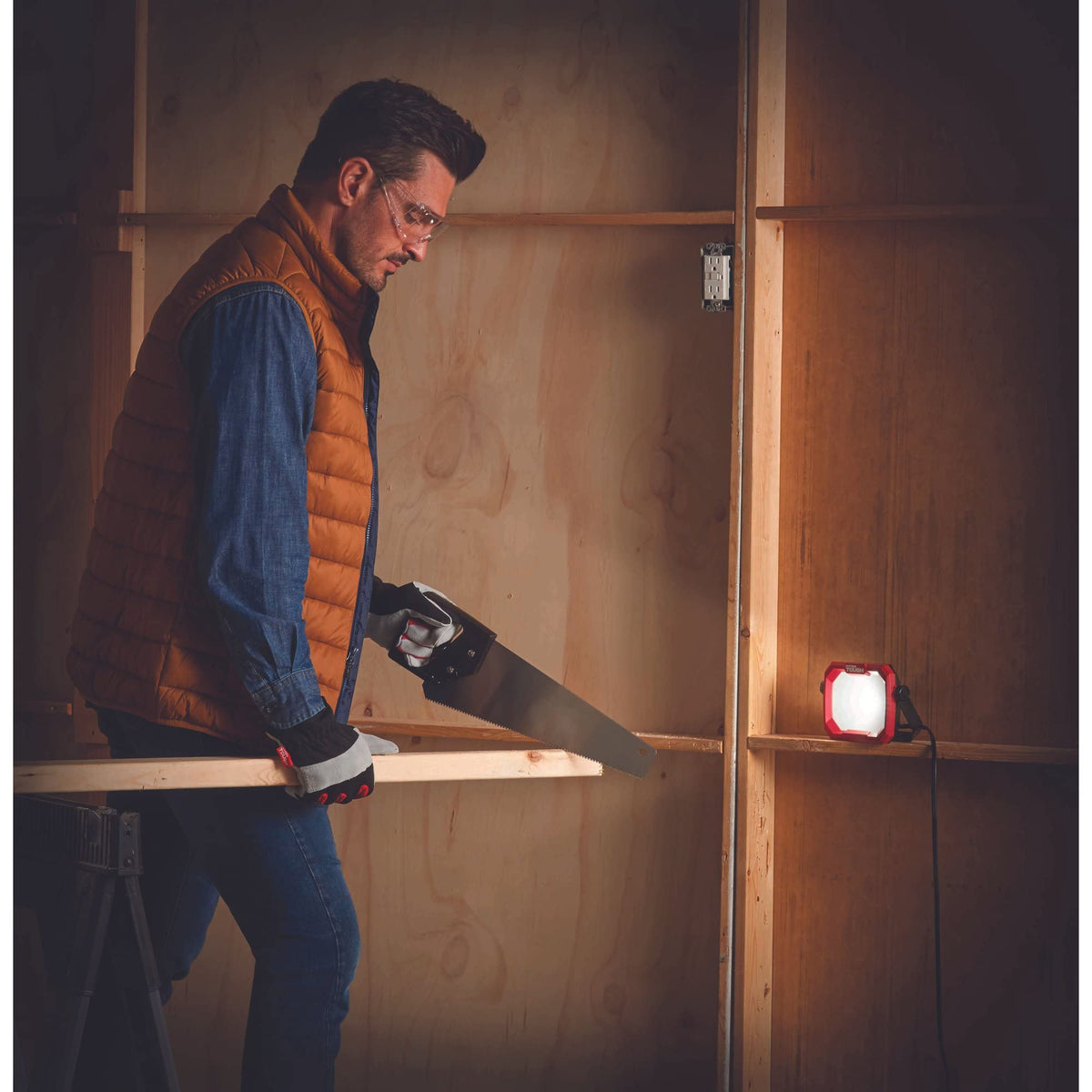 Man sawing a board in a workshop lit by the red LED work light on a shelf