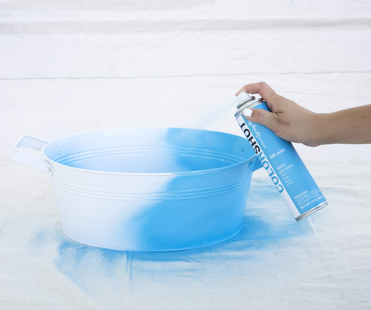 Hand spraying light blue paint onto a white metal tub on a drop cloth