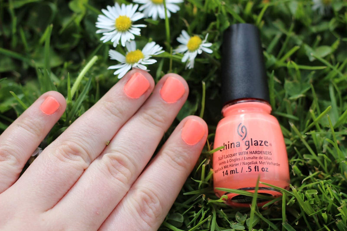 Hand with coral nails resting on grass beside the polish bottle and white daisies