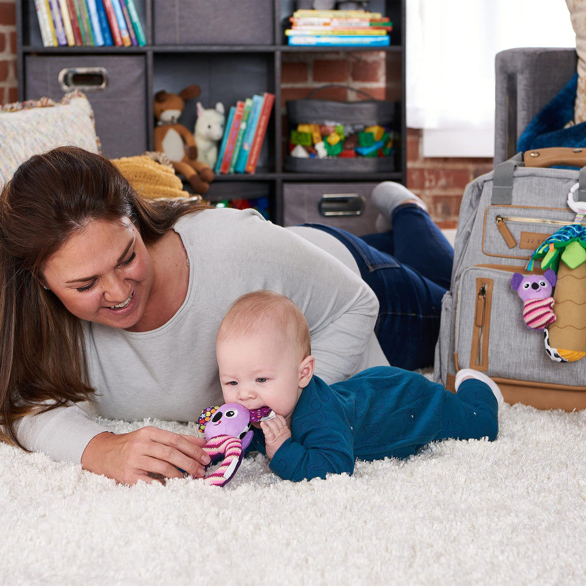 Baby on a rug chewing the purple Lamaze koala toy during tummy time with a parent