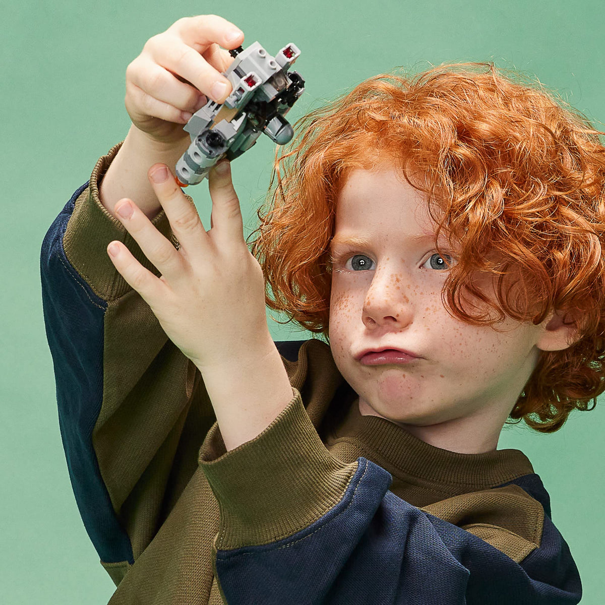 Red-haired child holding up the small grey Razor Crest build against a green wall