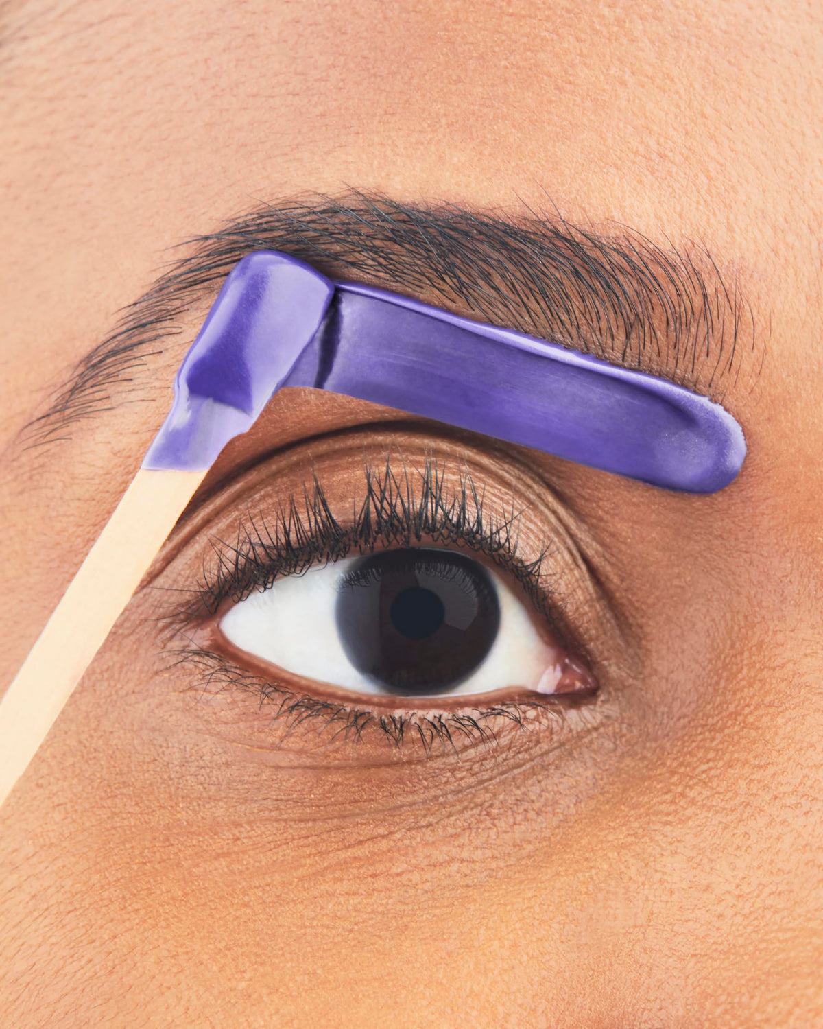 Close-up of melted purple wax applied along the lower edge of an eyebrow with a wooden applicator stick