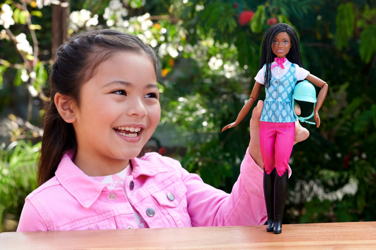 Smiling girl in a pink jacket holding up the Barbie Brooklyn riding doll outdoors