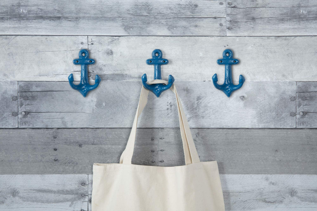 Three glossy blue anchor hooks on a grey wood wall, one holding a canvas tote