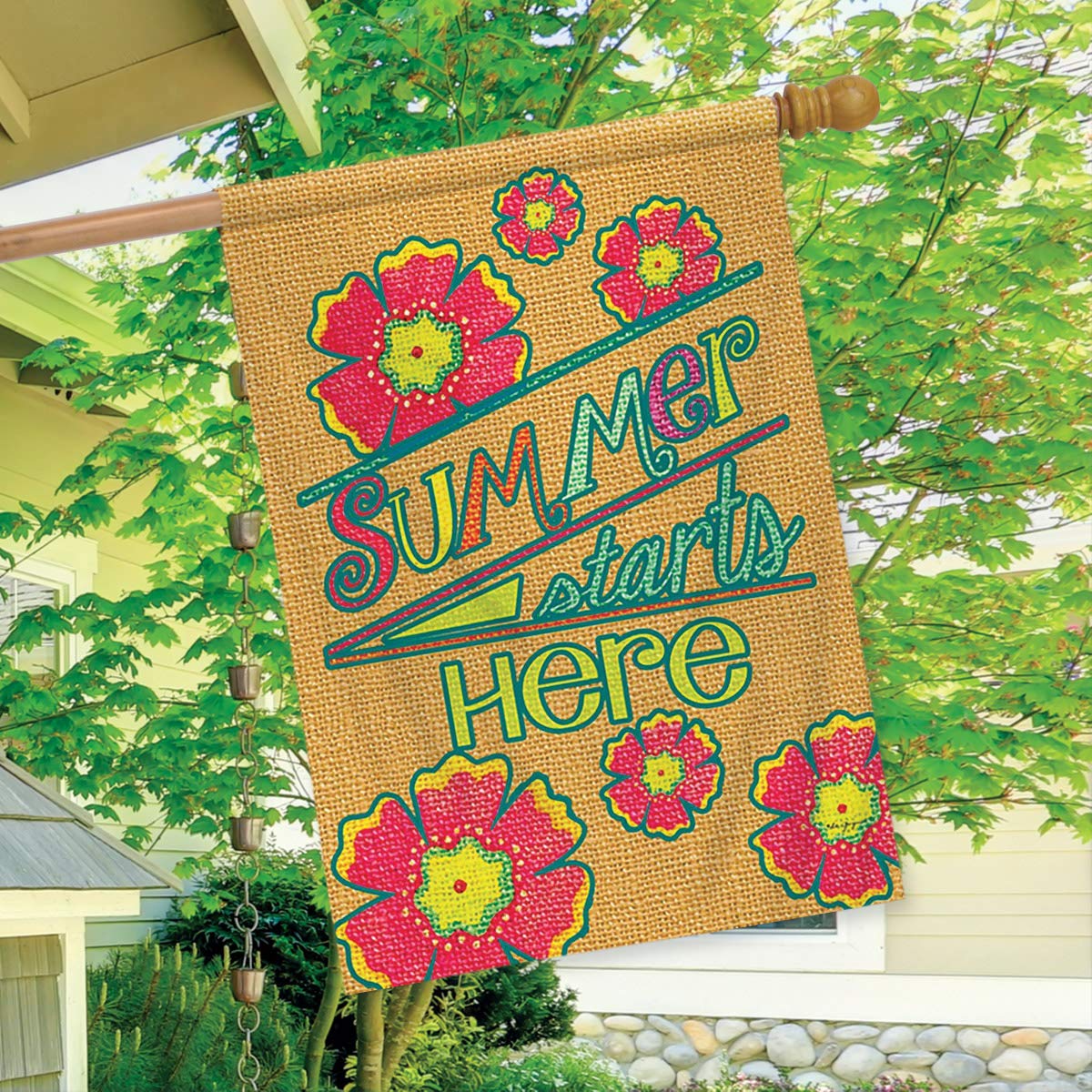 Summer starts here burlap-print flag hanging from a porch beside a leafy tree
