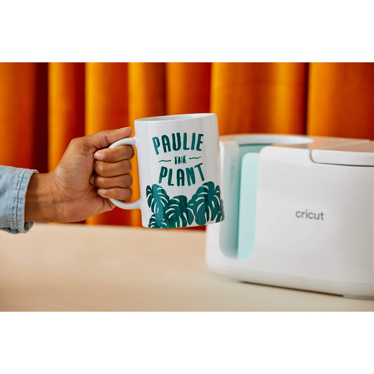 Hand holding a green Paulie the Plant mug next to the Cricut Mug Press