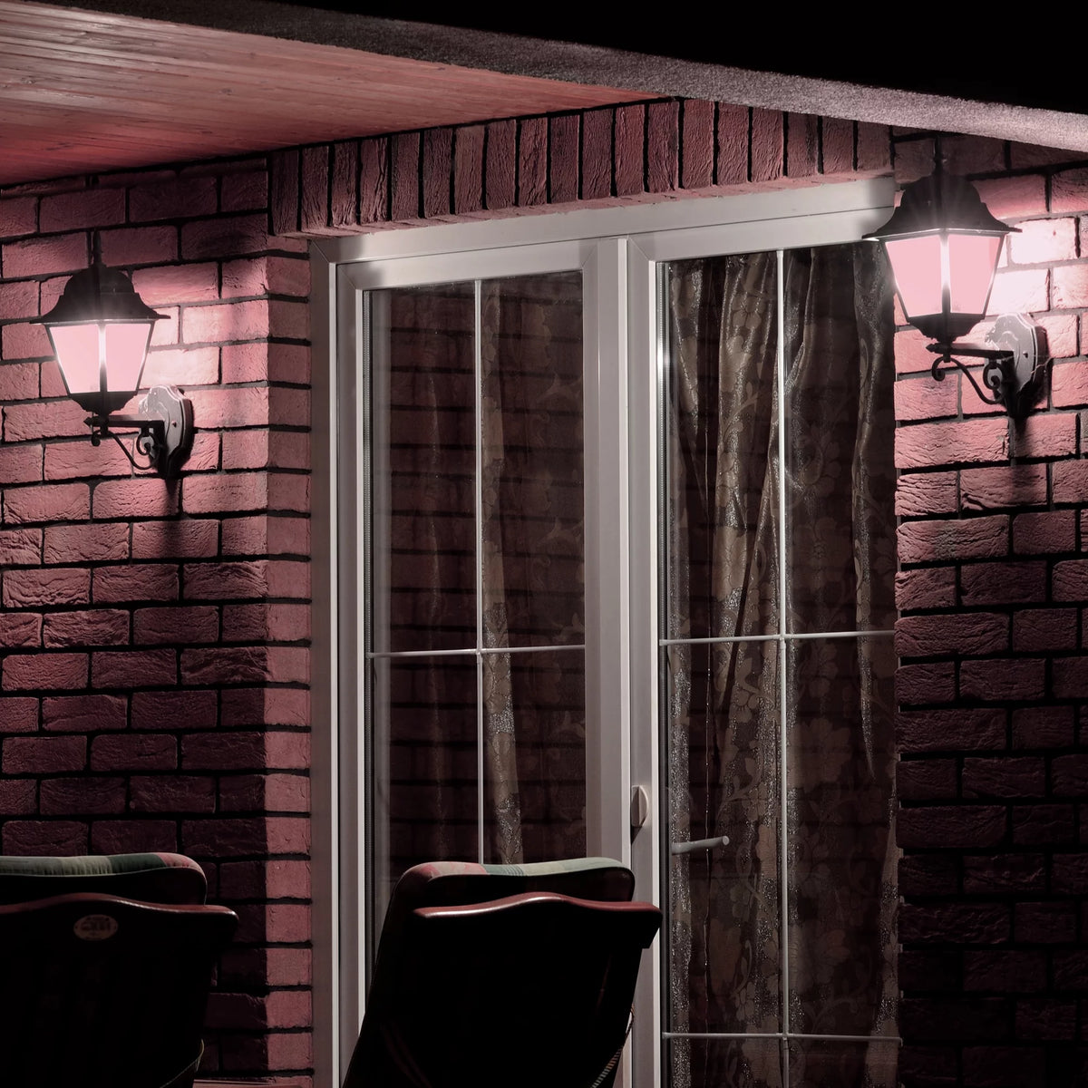 Two outdoor wall lanterns glowing pink beside patio doors on a brick wall