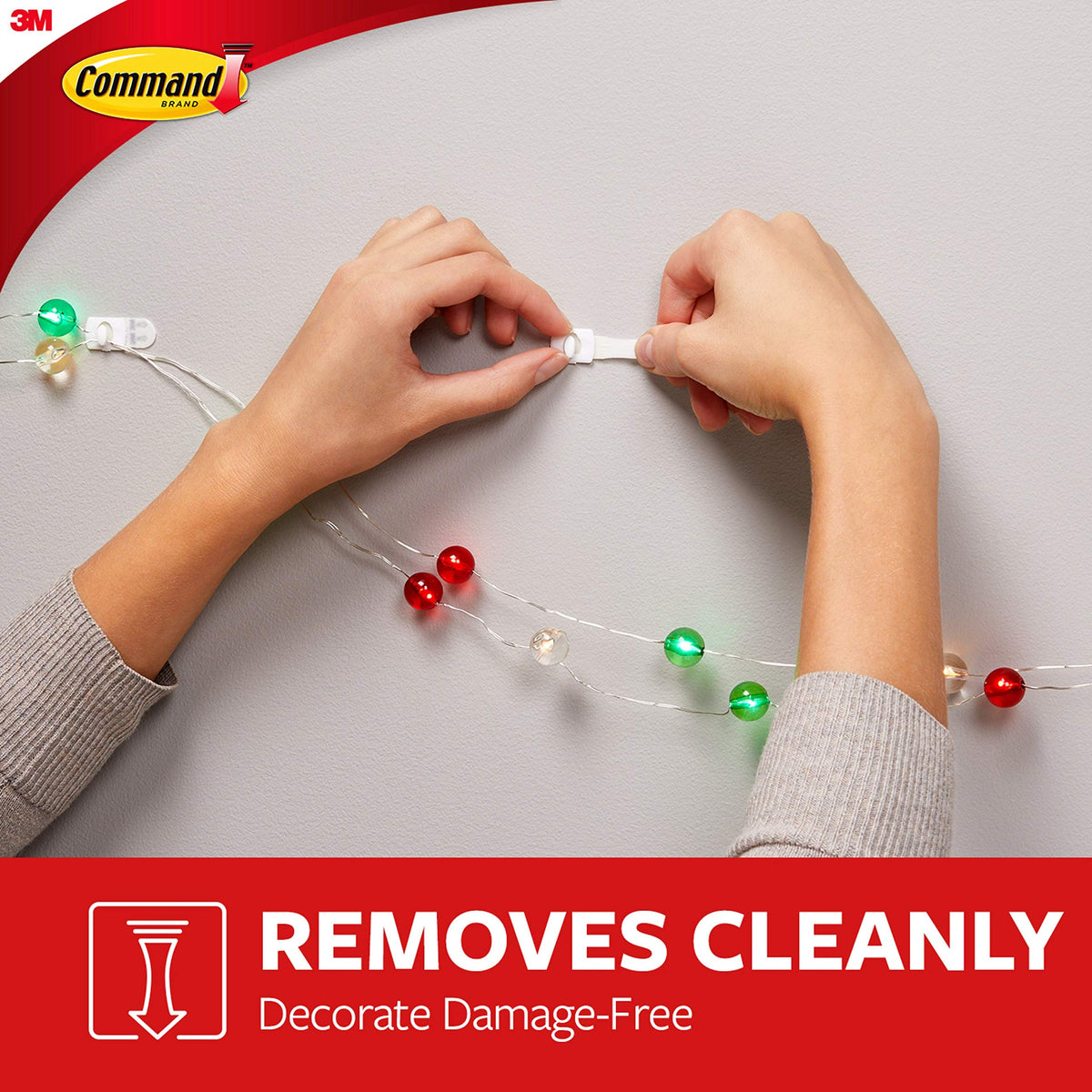 Hands stretching a clip's strip down a wall to remove it beside red and green string lights