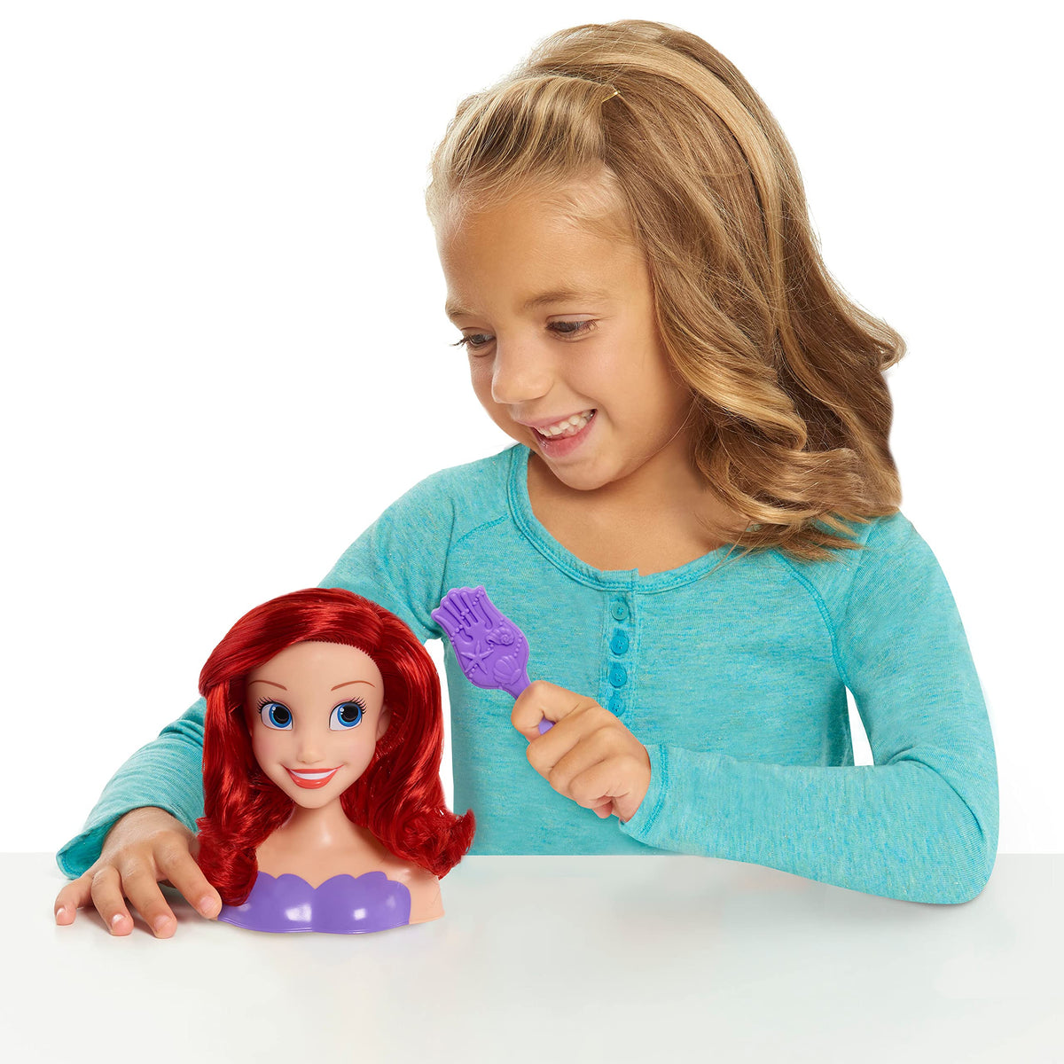 Smiling girl holding a purple seashell brush next to the red-haired Ariel styling head