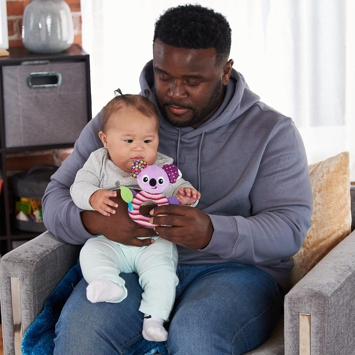 Parent on an armchair showing a purple Lamaze koala rattle to a baby on their lap