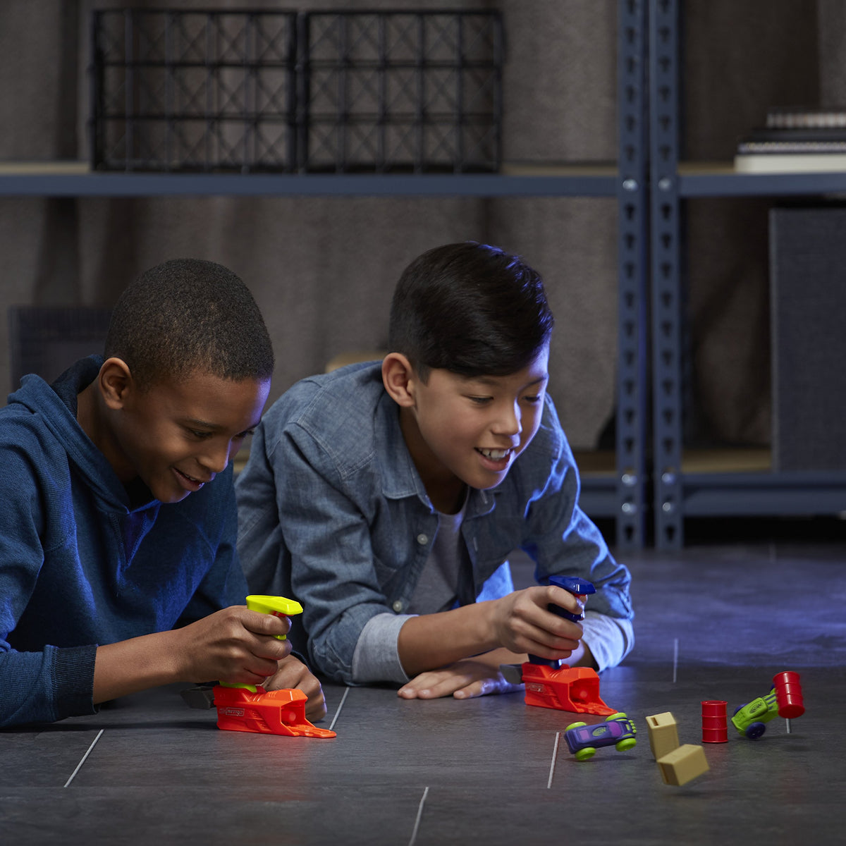 Two boys launching foam cars into red barrels and tan crates on a tiled floor