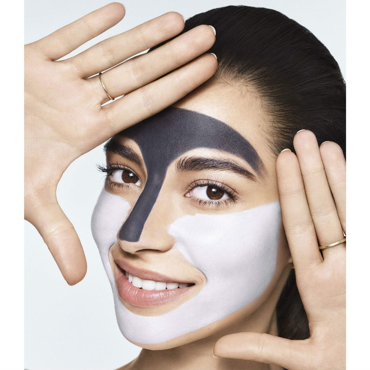 Woman framing her face with her hands, wearing dark and white clay masks on different zones