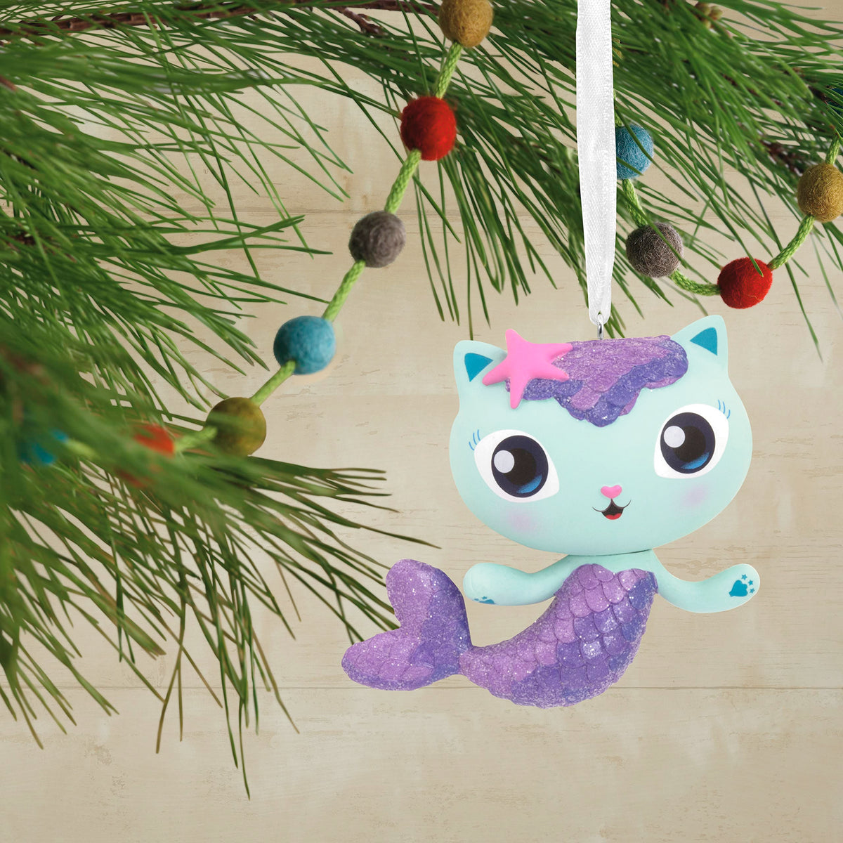 Mermaid cat ornament hanging on a pine branch with a felt ball garland