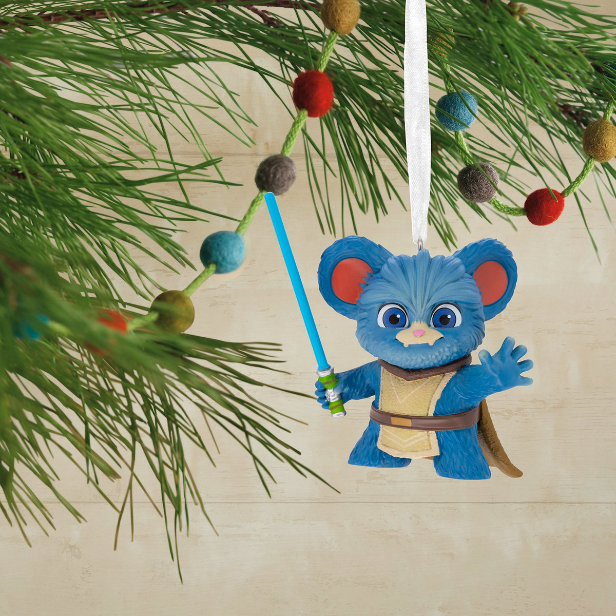 Blue fuzzy creature ornament hanging on a pine branch with a felt ball garland