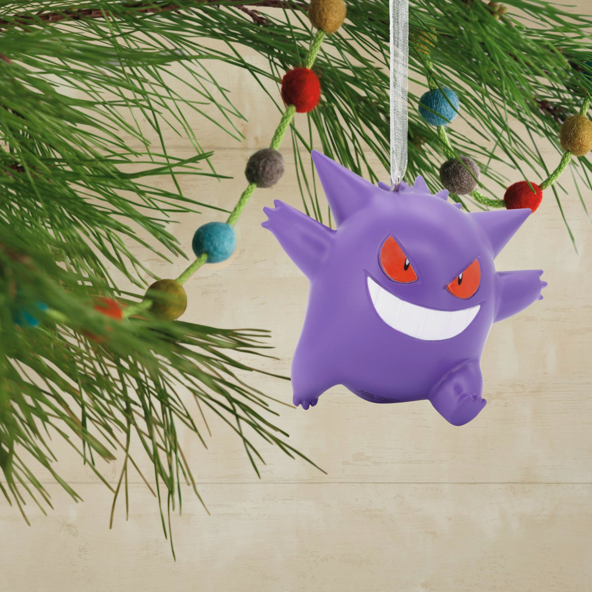 Purple grinning ghost ornament hanging on a pine branch with a felt ball garland