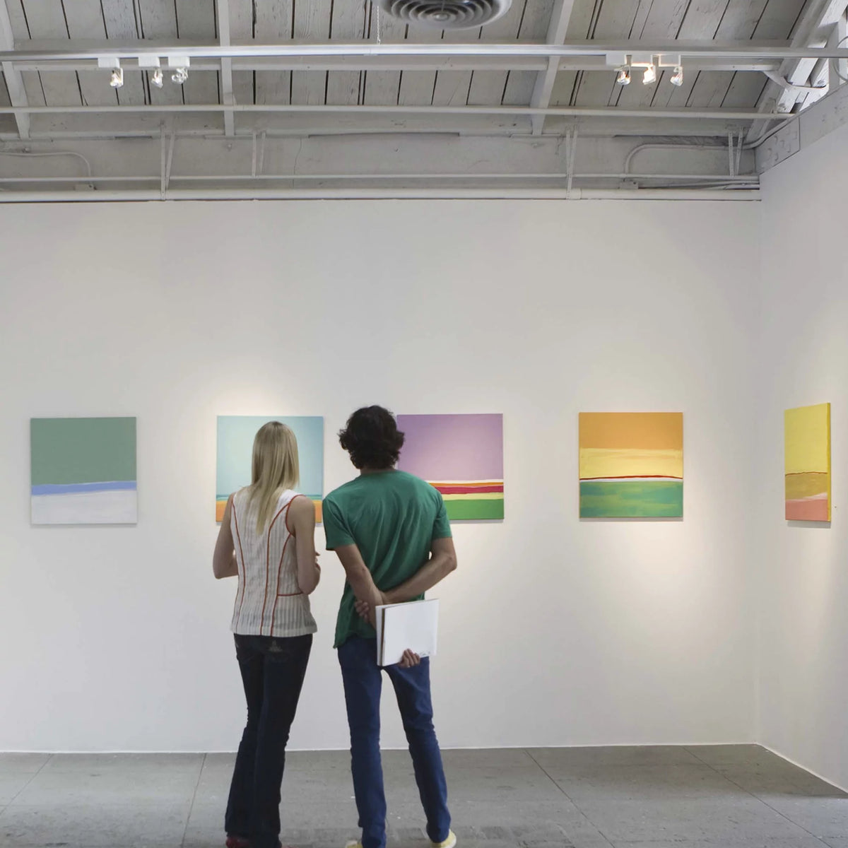 Two people viewing colourful paintings lit by track spotlights in a white gallery