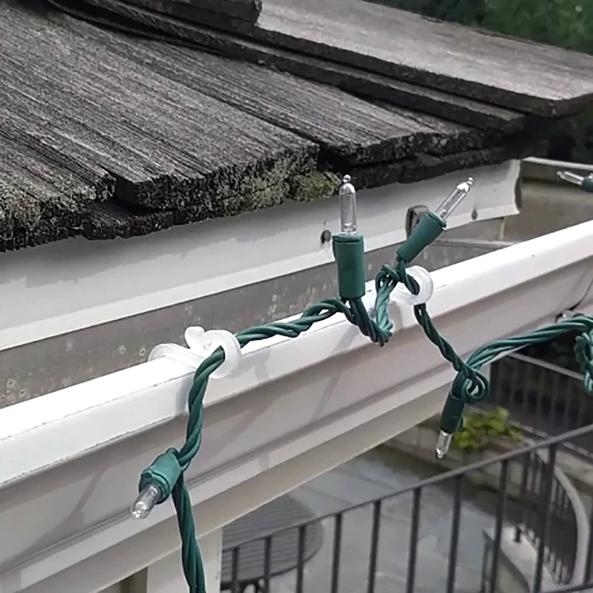 Clear hooks clipping green-wire mini string lights onto a gutter under wood shingles