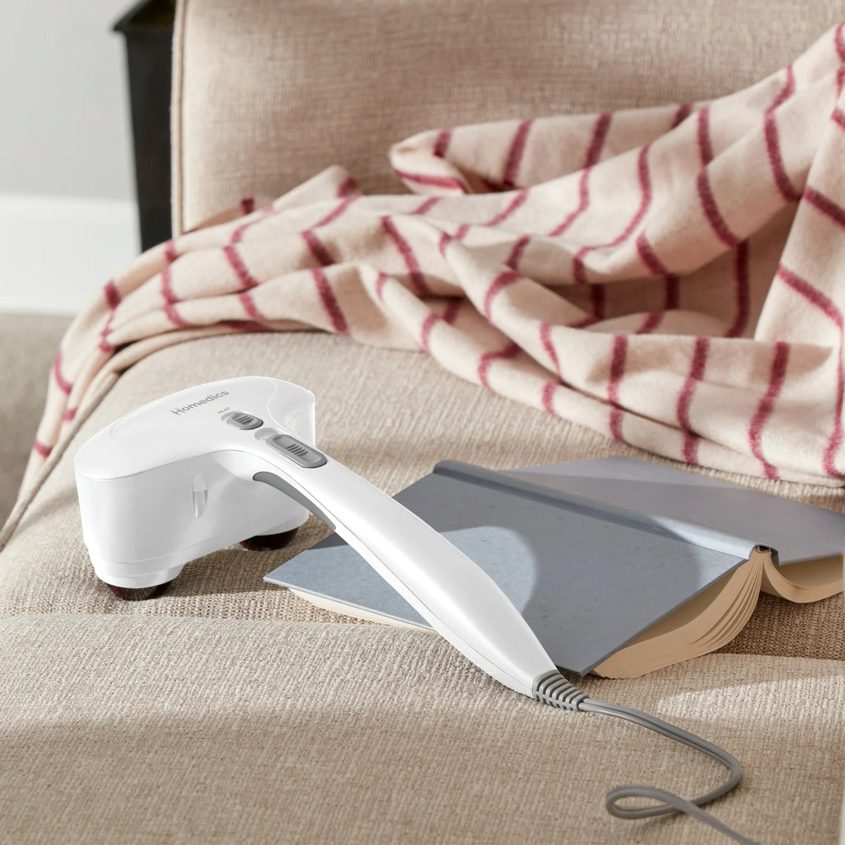 White handheld massager with heat and speed switches resting on a sofa by a book