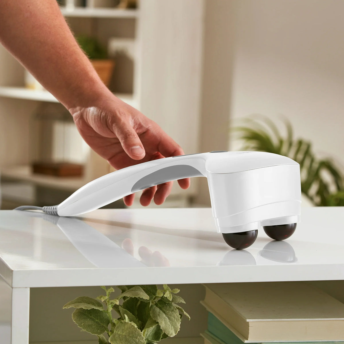 Hand reaching for the white massager resting on its two dark nodes on a white table
