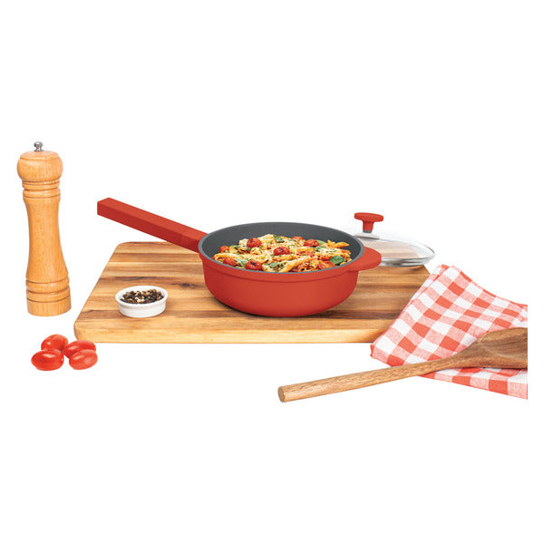 Red 8 inch pan filled with pasta on a wooden board, glass lid, pepper mill and checked towel beside it