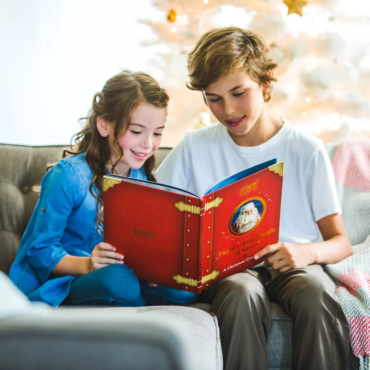 Two children on a sofa reading the red Santa storybook in front of a white Christmas tree