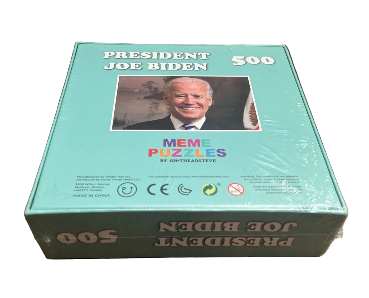 A boxed puzzle titled "PRESIDENT JOE BIDEN 500" sits on a wooden tabletop. The box is light aqua/turquoise with a photo of Joe Biden on the front, the text "MEME PUZZLES BY SHTHEAISTEV8" in the center, and the product name printed upside down on the lower box.