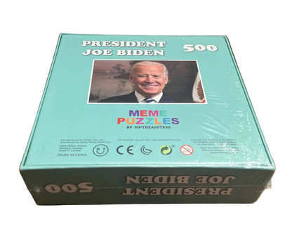 A boxed puzzle titled "PRESIDENT JOE BIDEN 500" sits on a wooden tabletop. The box is light aqua/turquoise with a photo of Joe Biden on the front, the text "MEME PUZZLES BY SHTHEAISTEV8" in the center, and the product name printed upside down on the lower box.