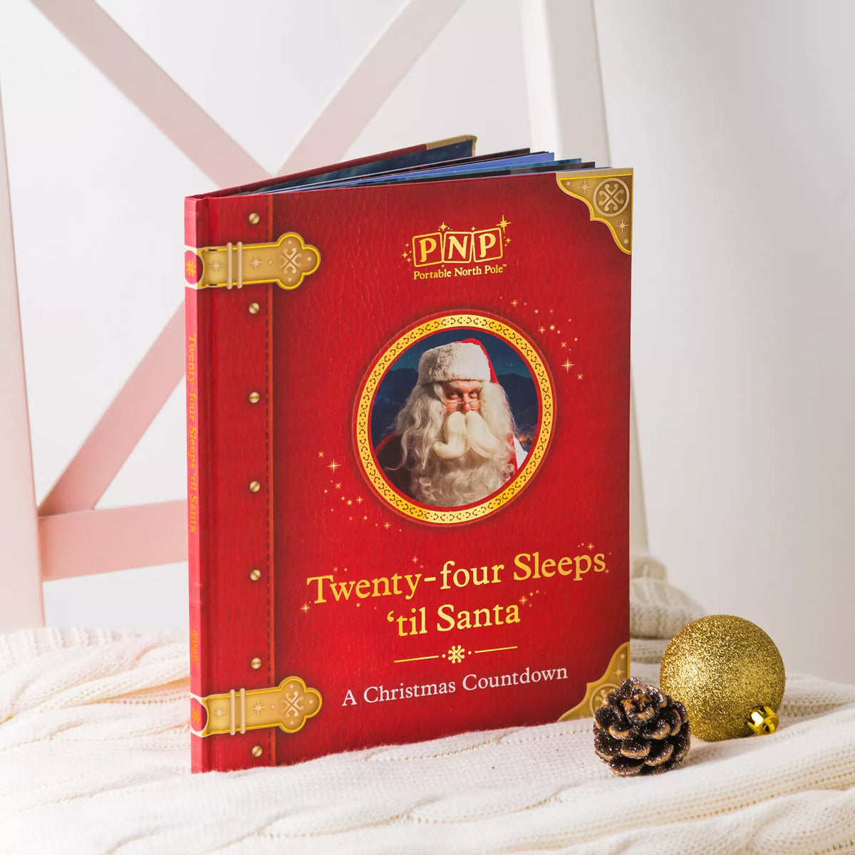 Red Santa storybook standing on a knit throw beside a pine cone and gold bauble