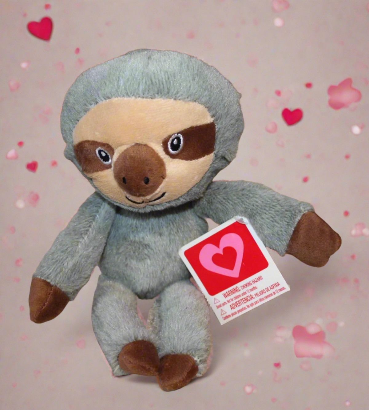 Grey plush sloth with a heart tag on a pink background scattered with hearts