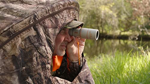 Man in camo gear looking through the scope by a pond