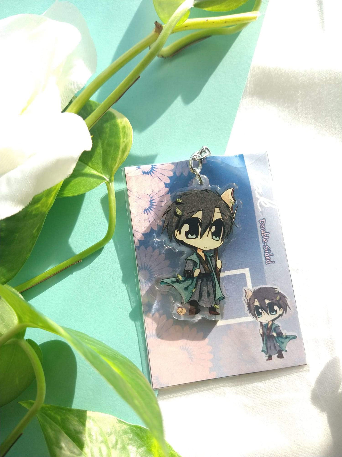 Chibi Hak acrylic keychain on its blue flower backing card beside green leaves