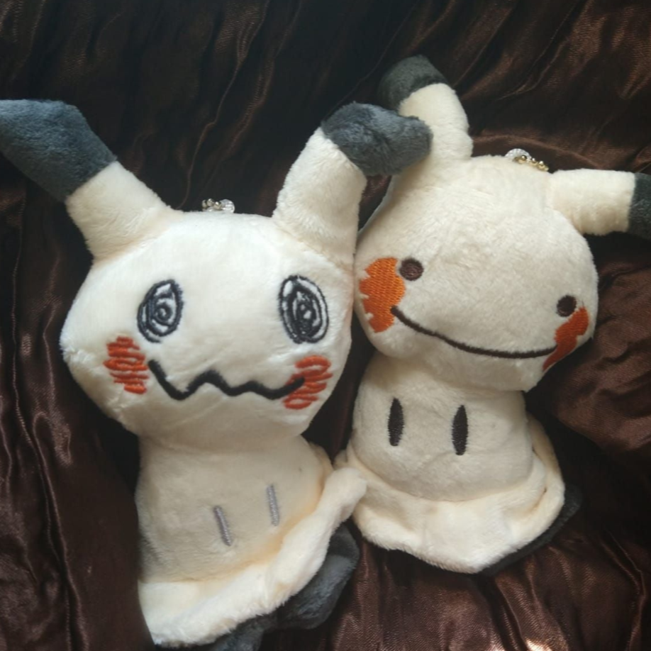 Two cream Mimikyu keychain plushes with embroidered faces on brown satin