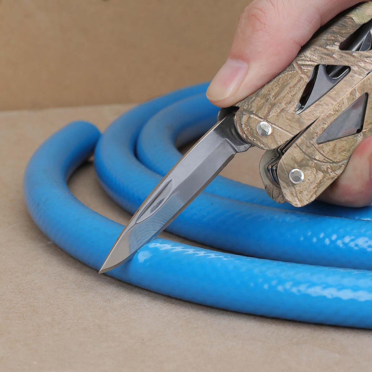Camo multi-tool knife blade slicing a blue rubber hose